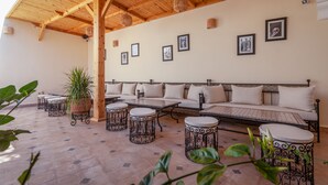 Un área de estar al aire libre techada, con techo de madera, bancos acolchados, mesas redondas y plantas en macetas.