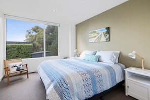 A bedroom with a large window, a bed with patterned bedding, a bedside table with a lamp, and a painting on the wall. - Seasoon (Lorne)