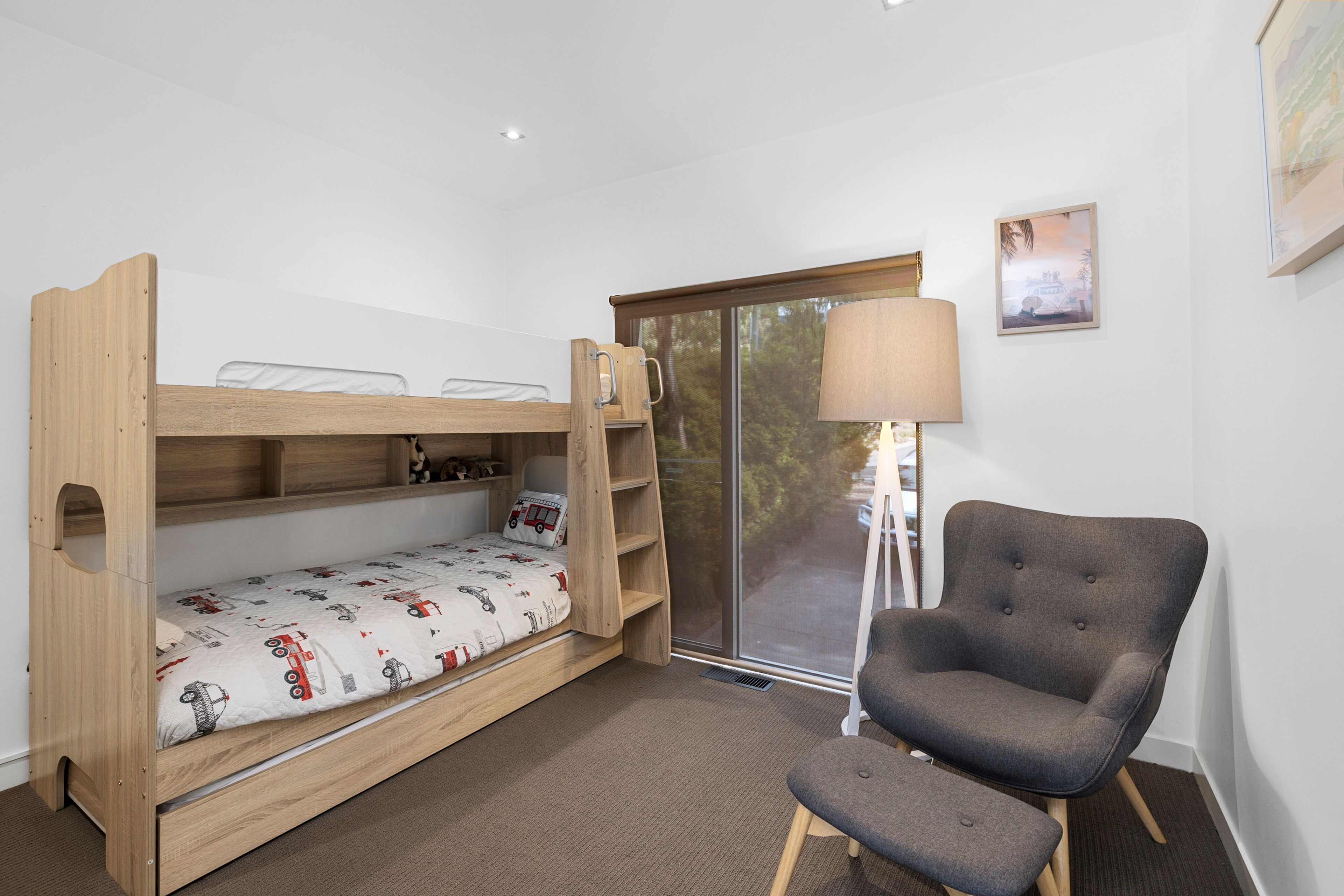 Un lit superposé avec un bureau escamotable, un fauteuil gris, une lampe sur pied et une fenêtre donnant sur les arbres.