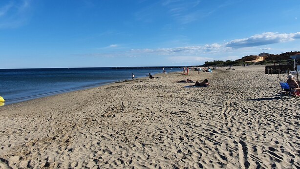 Ein sandiger Strand mit blauem Himmel, wenigen Personen und Gebäuden im Hintergrund.