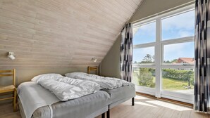 Ein Schlafzimmer mit schrägverdeckter Decke, einem großen Fenster mit Vorhängen, einem Bett mit weißer Bettwäsche und Holzboden.