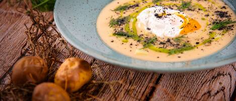 A bowl of soup with a poached egg, garnished with herbs and black pepper, on a wooden table with dried plants and potatoes.