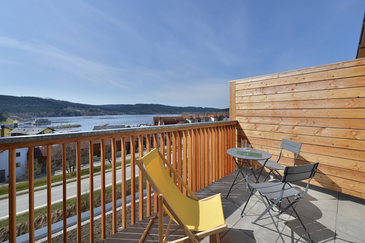 Un balcon avec vue sur un lac et des bâtiments, une balustrade en bois et du mobilier d’extérieur.