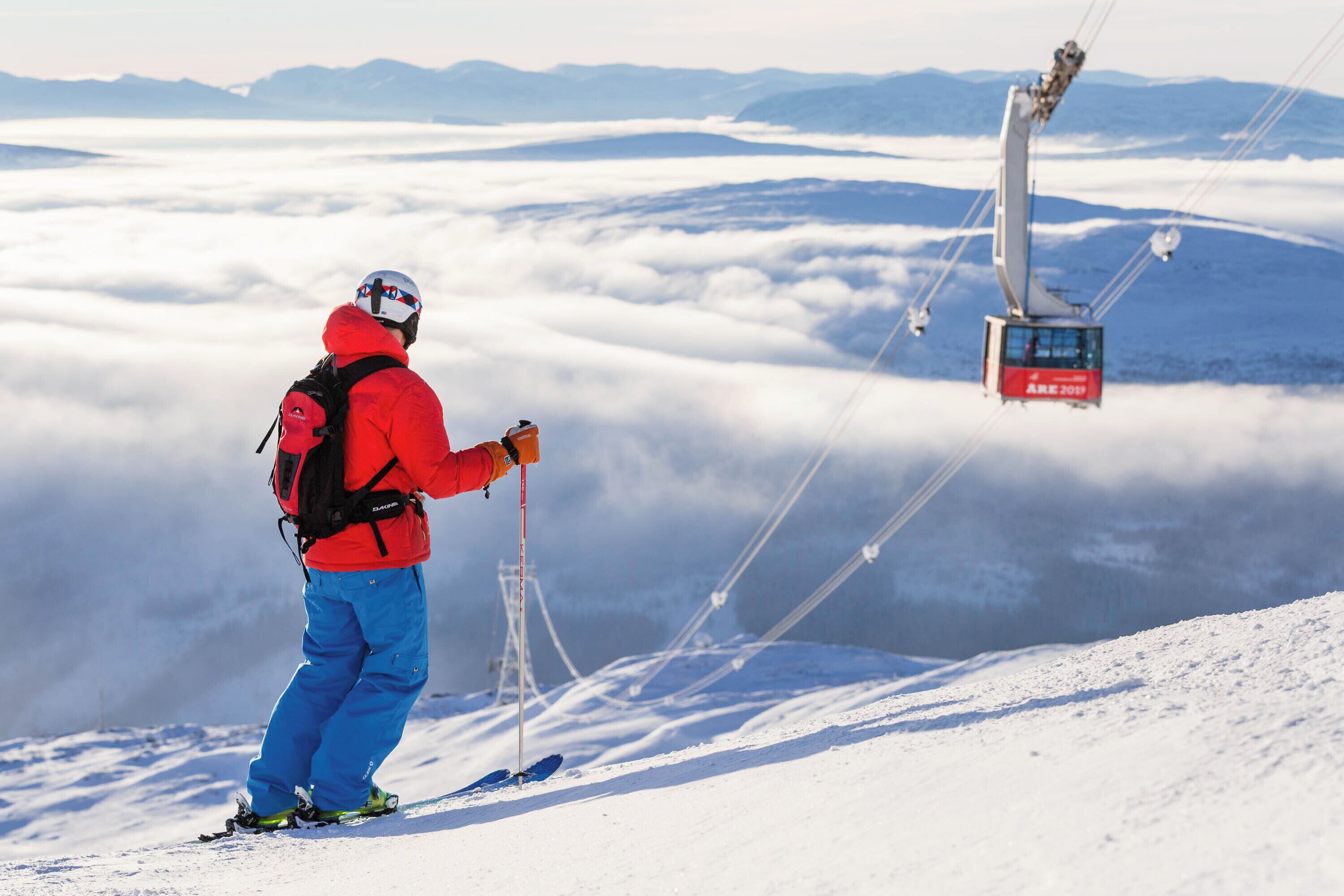 A person in red and blue ski gear standing on a snowy slope with a cable car in the background.
