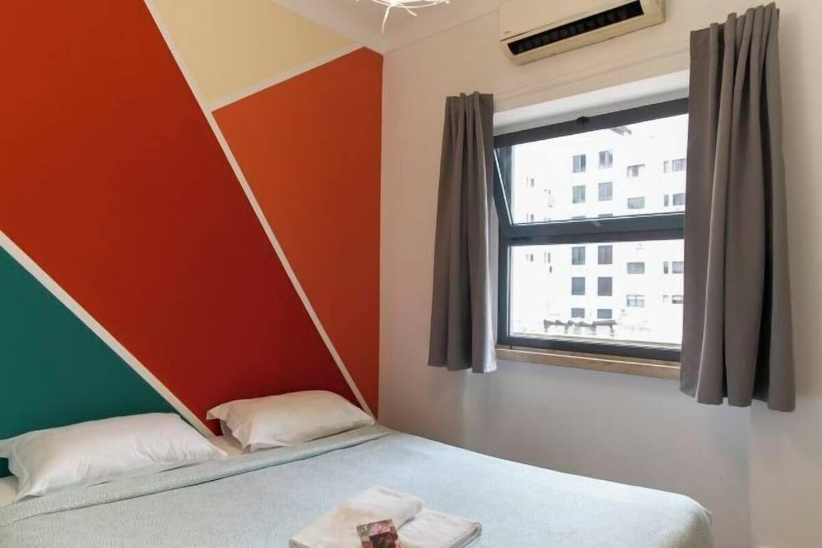 Uma cama com almofadas, uma janela com cortinas e vista para edifícios lá fora.
