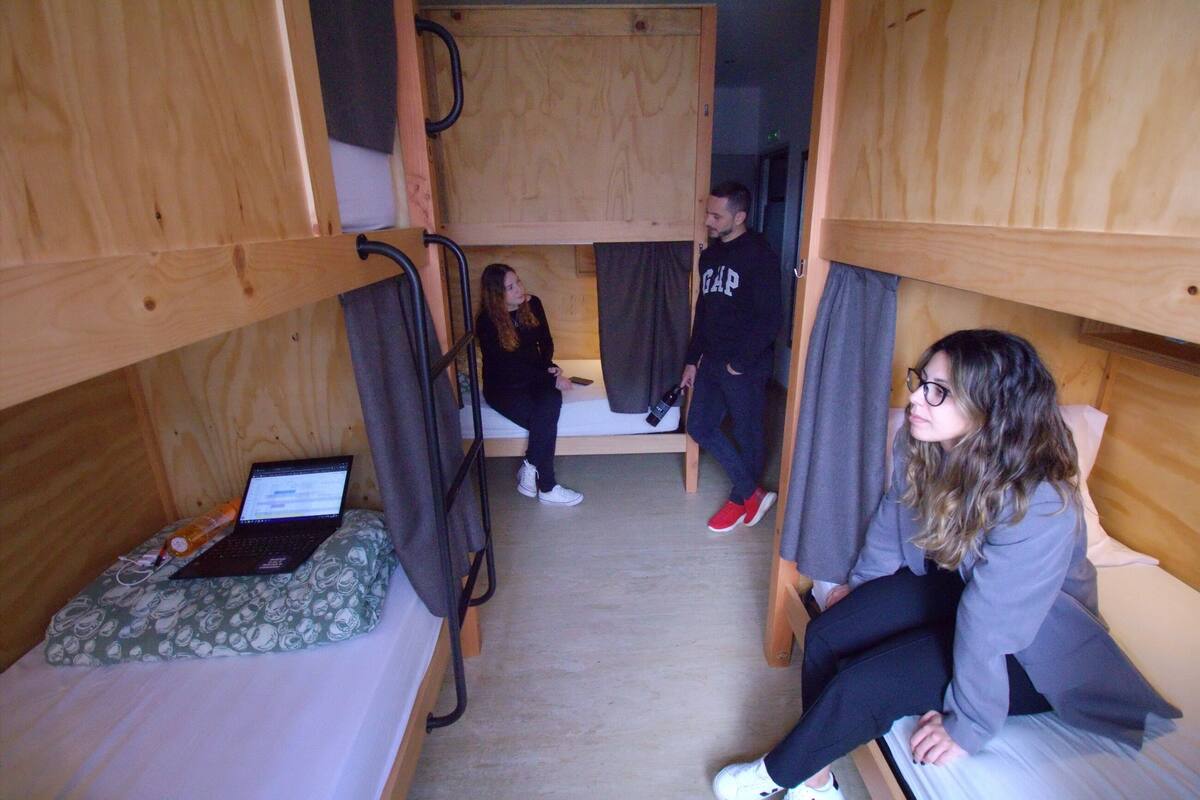 Quarto tipo dormitório com beliches, um laptop sobre um travesseiro, e uma pessoa sentada na cama.