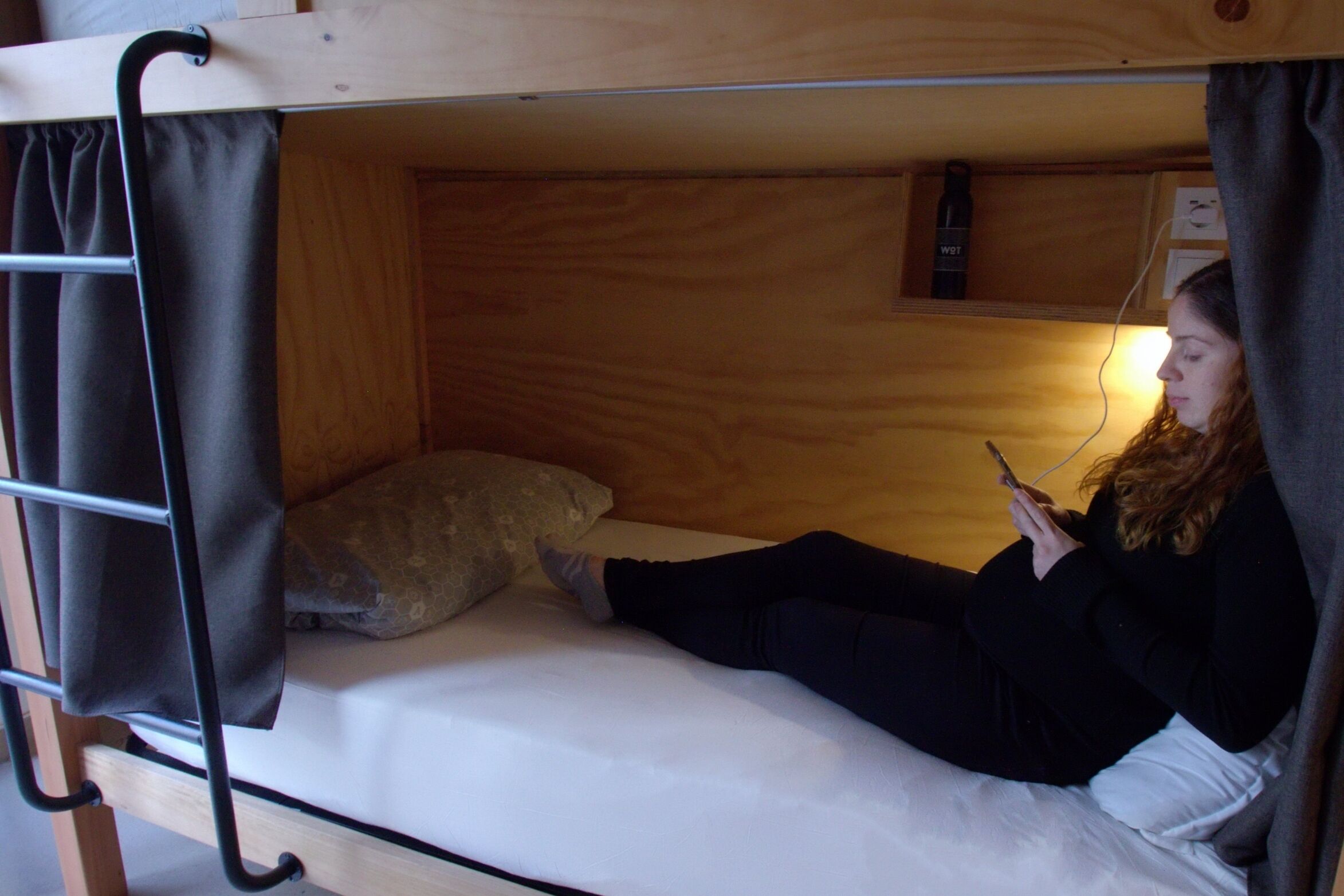 A bunk bed with a person lying on the lower bunk, a pillow, and a small shelf with a bottle.