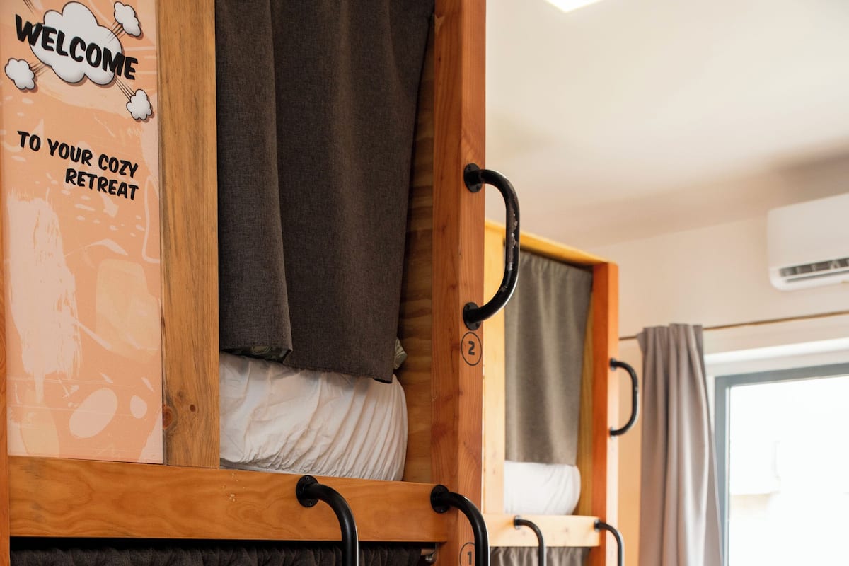 Quarto com beliches, um cartaz de boas-vindas e uma janela com cortinas.