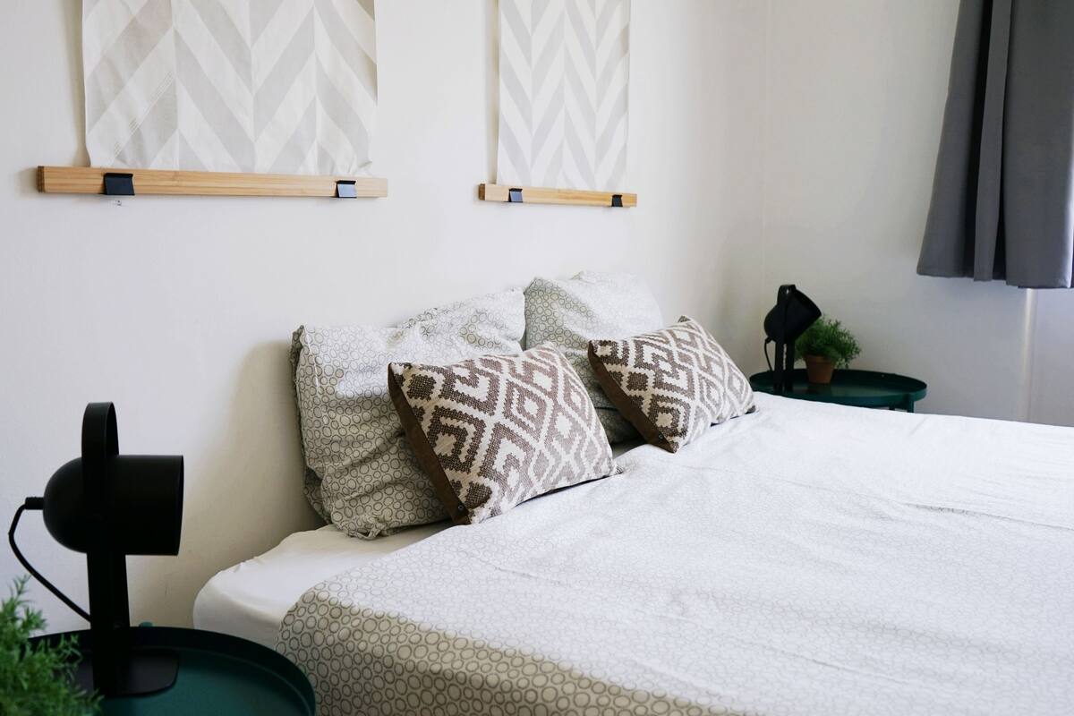 Uma cama bem arrumada com almofadas estampadas, uma luminária de cabeceira e um ponto de luz fixado na parede.