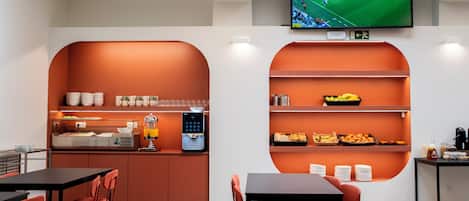 Un área de cocina moderna con un televisor de pantalla plana mostrando un evento deportivo, una cafetera y una barra de desayuno con diferentes alimentos.