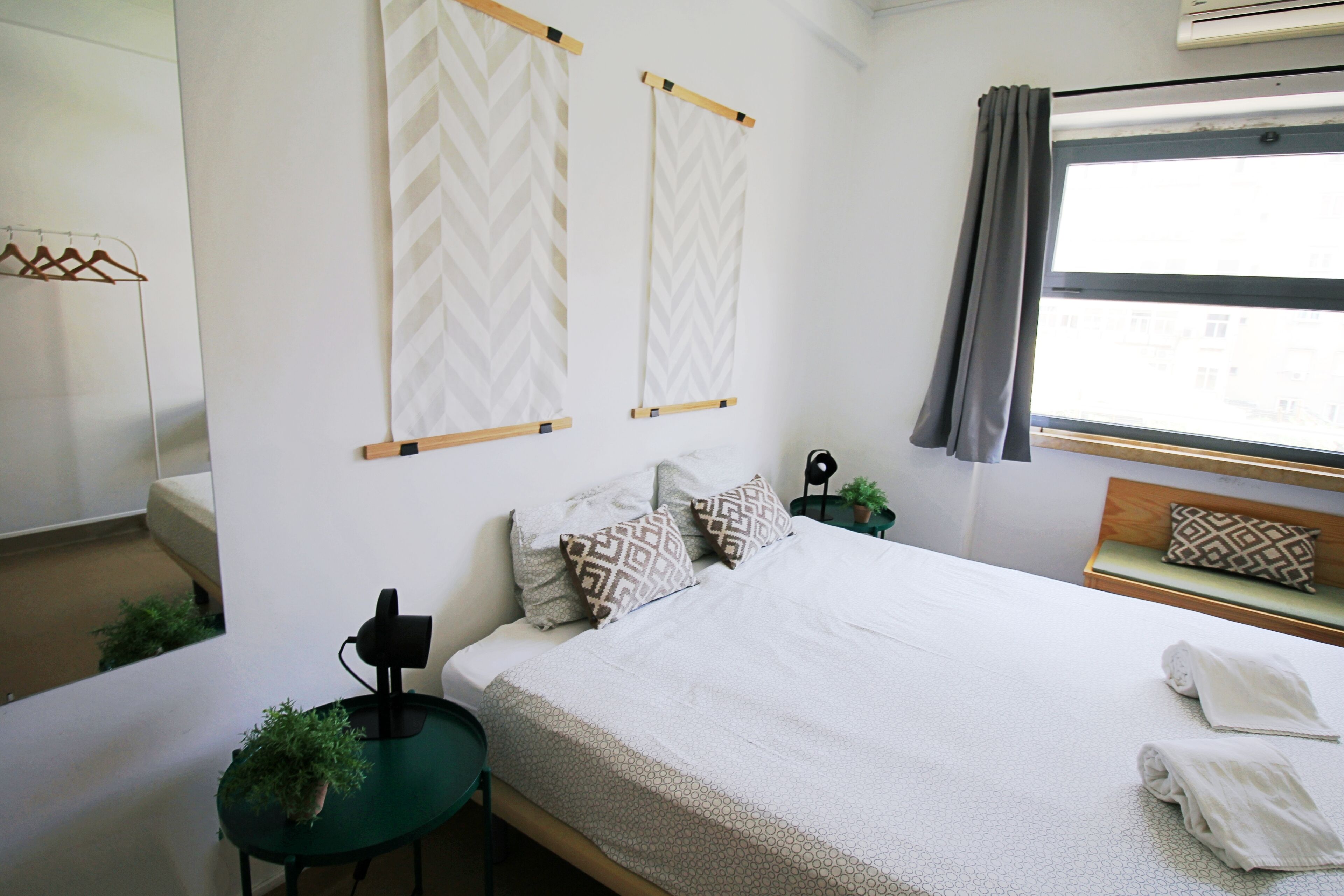 A neatly arranged bedroom with a bed, pillows, a bedside table with a plant, a small table with a lamp, and a window with curtains.