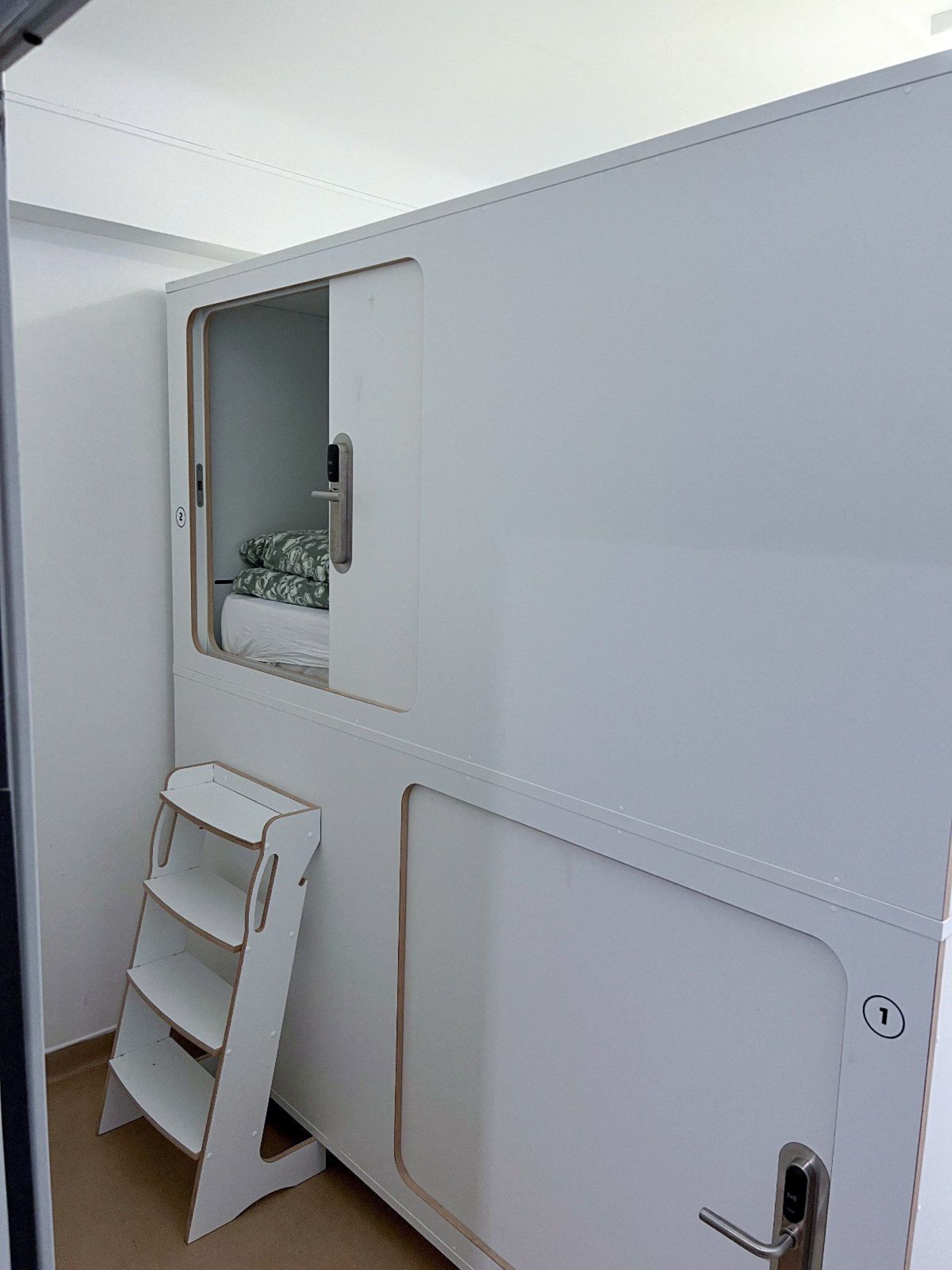 Una habitación con cama, ventana, puerta y un pequeño estante blanco.