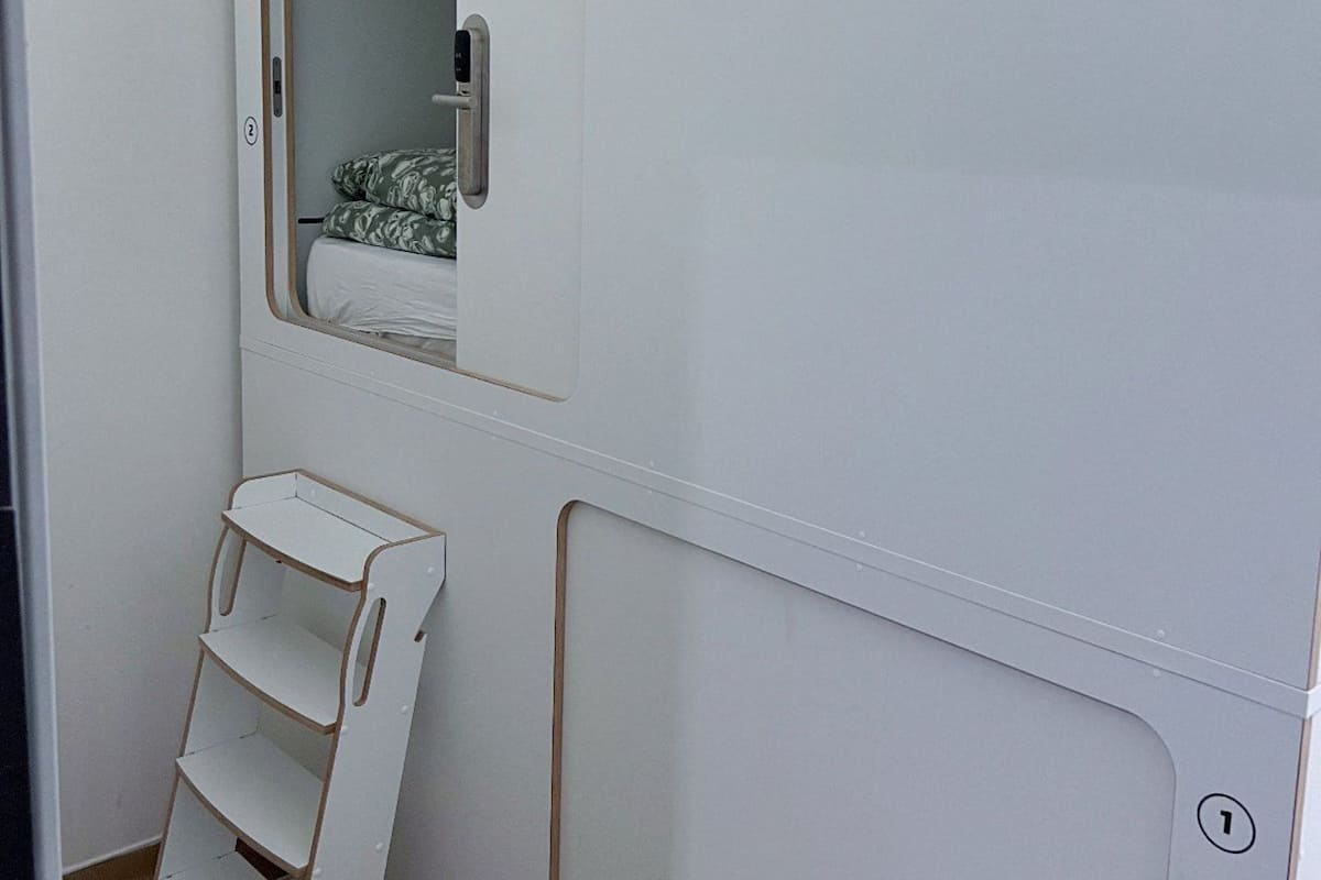 Um quarto com uma cama, uma janela, uma porta e uma pequena estante branca.