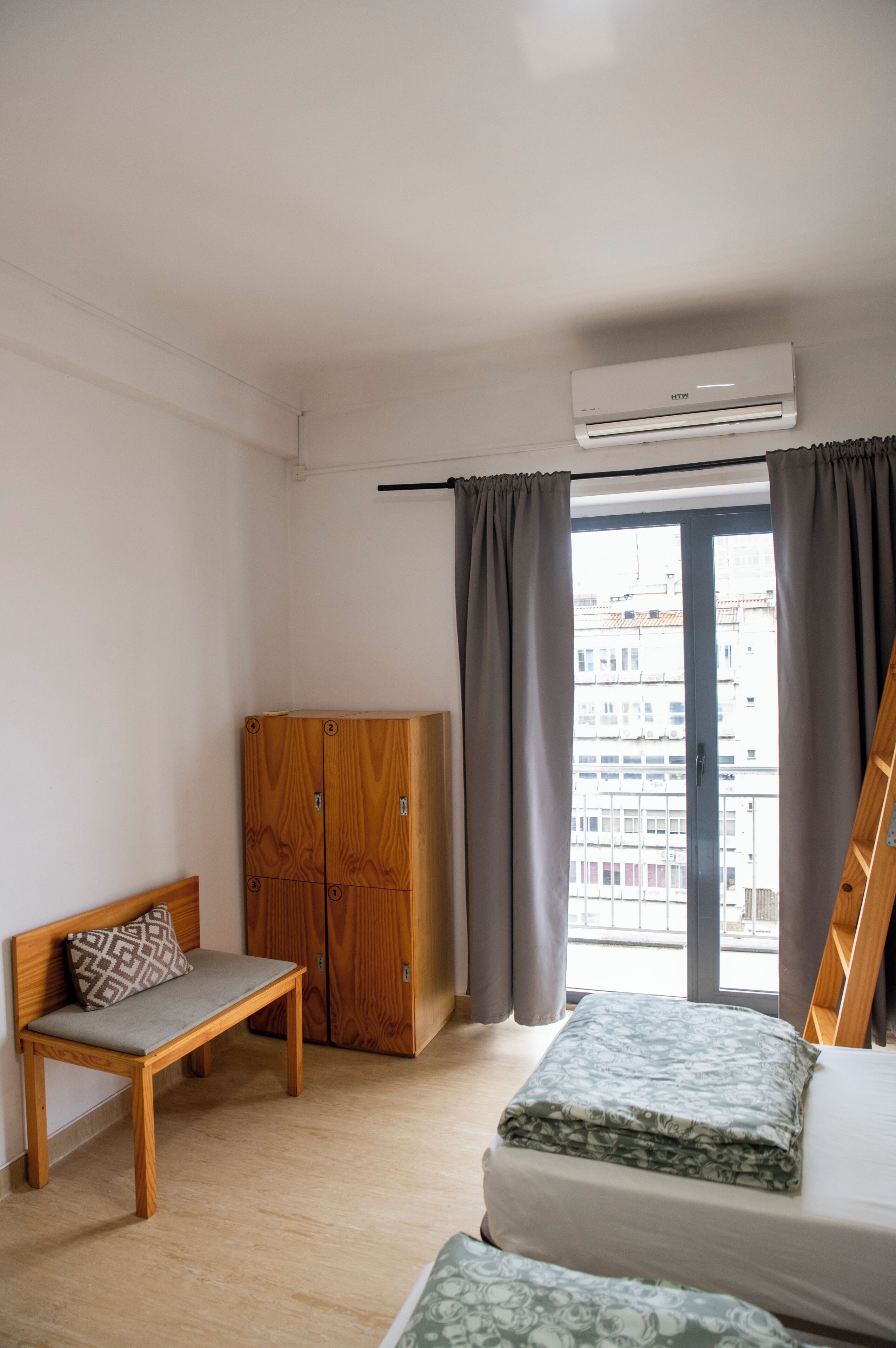 Quarto pequeno e limpo com um banco de madeira, guarda-roupa, cama com manta estampada e aparelho de ar condicionado.
