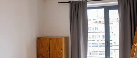 Quarto pequeno e limpo com um banco de madeira, guarda-roupa, cama com manta estampada e aparelho de ar condicionado.