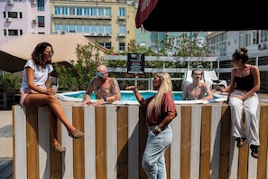A group of people sitting by a poolside with a wooden fence and a signboard in the background. - WOT Nomad Social (Lisbon)