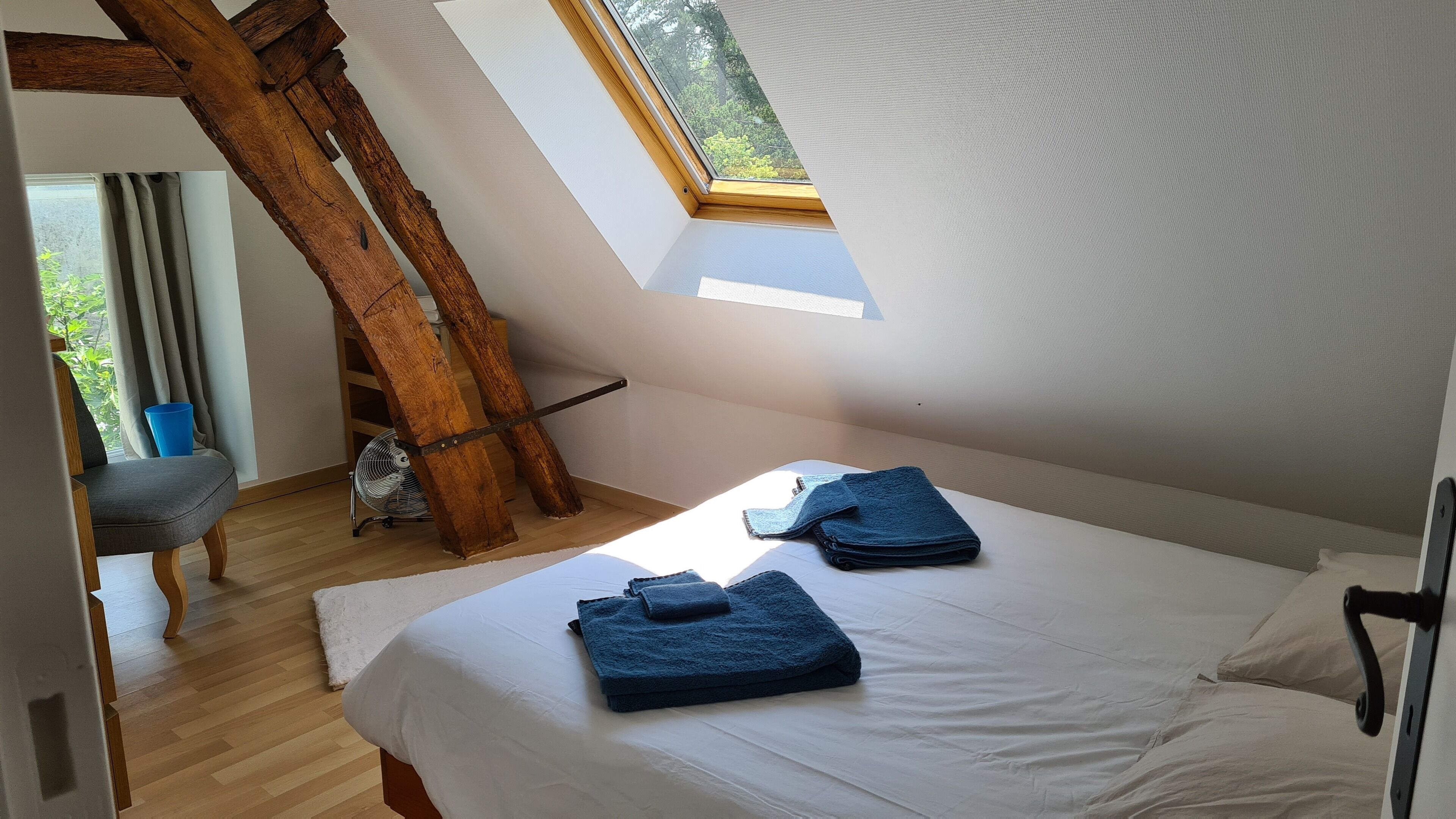 Un dormitorio con techo inclinado, tragaluz, cama con sábanas blancas y toalla azul.