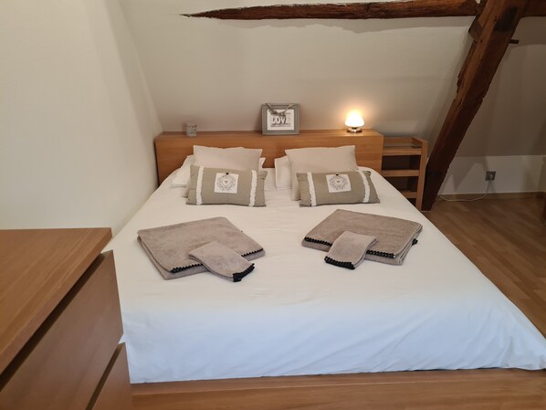 Ein ordentlich bezogenes Bett mit weißen Leinentüchern, zwei beigen Handtüchern und einem gerahmten Bild am Kopfteil.