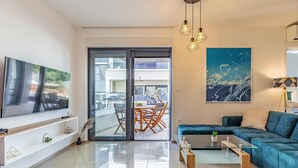 Ein modernes Wohnzimmer mit einem Flachbildfernseher, einem Glastisch, einem blauen Sofa und einem Essbereich mit Tisch und Stühlen.