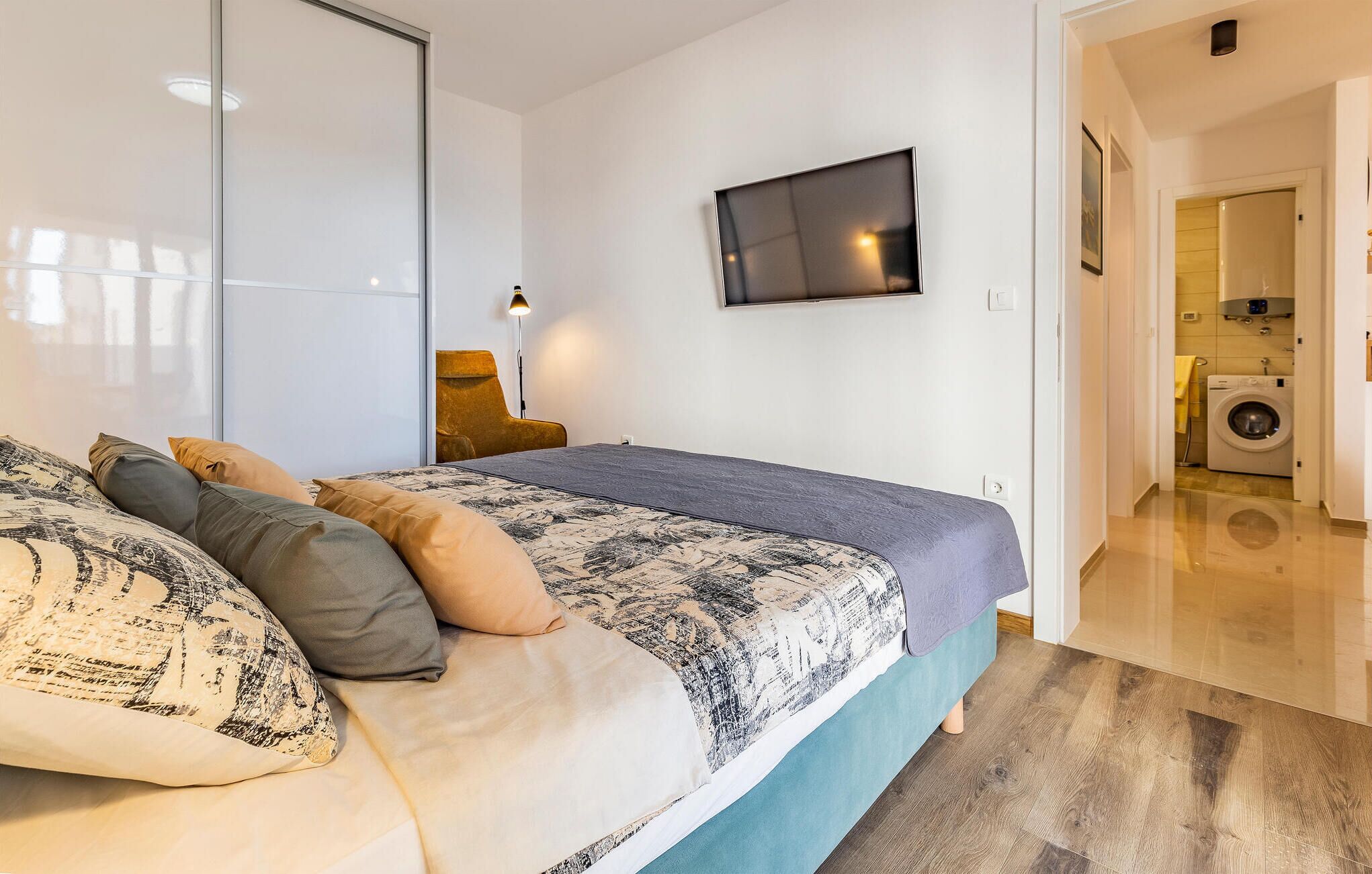 A modern bedroom with a bed, a TV mounted on the wall, and a washing machine in the adjacent room.