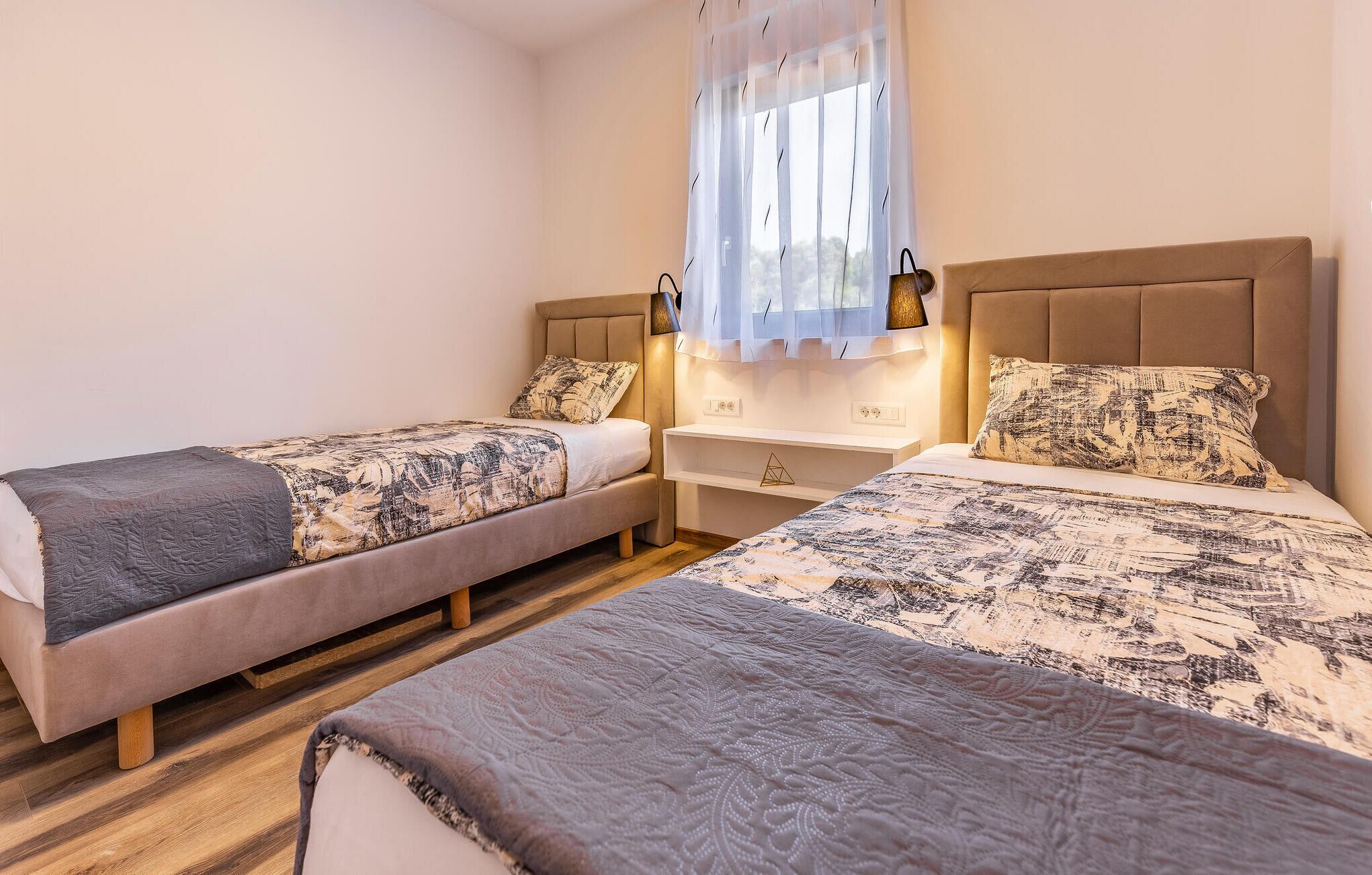 A bedroom with two beds, a nightstand, a lamp, and a window with a sheer curtain.