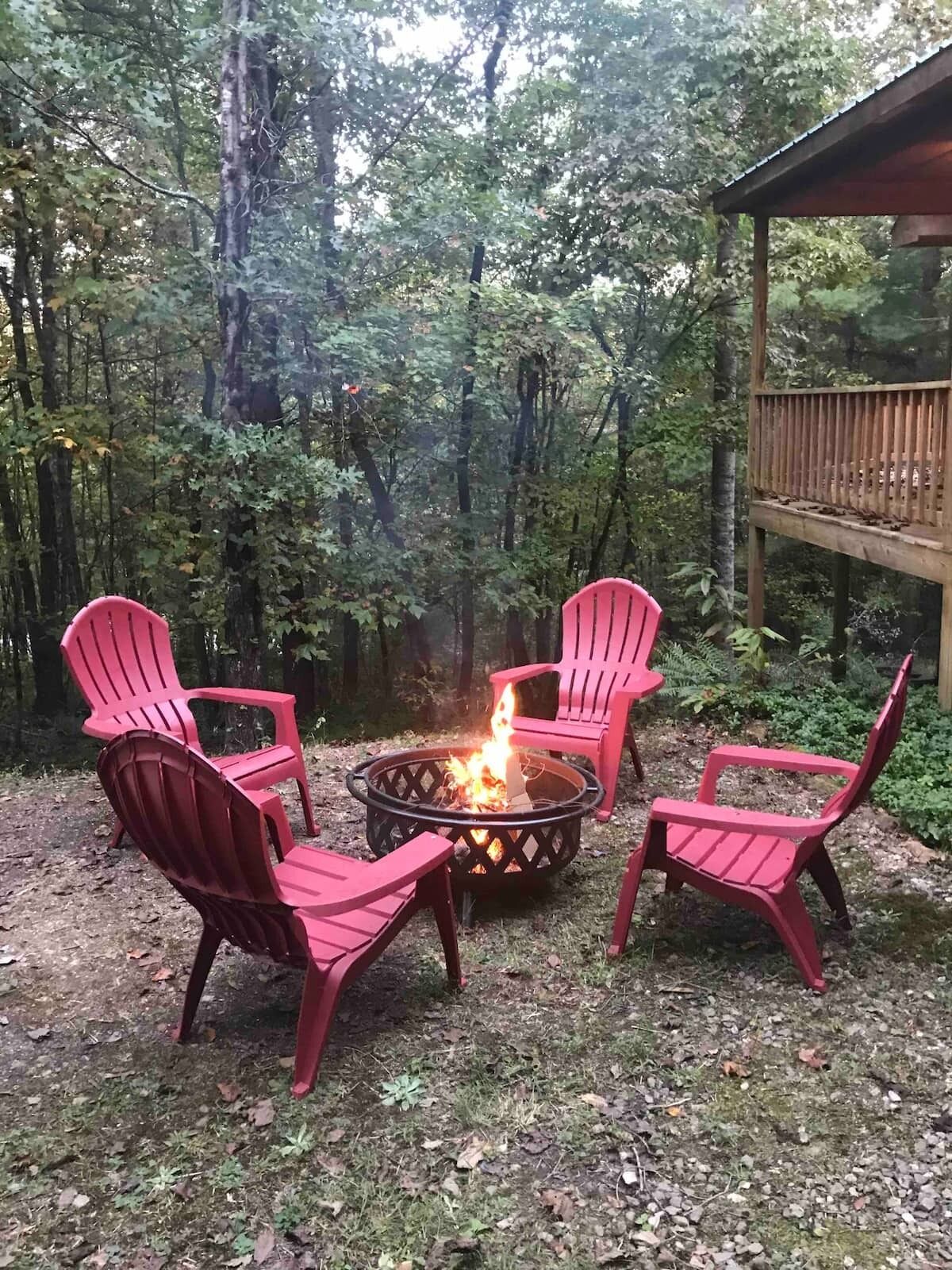 A fire pit with four Adirondack chairs arranged around it, set in a forested area with a wooden deck in the background.