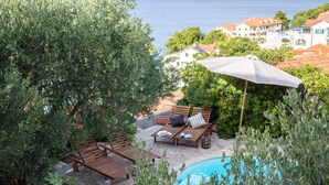 A poolside area with wooden lounge chairs and a large umbrella, surrounded by greenery and overlooking a coastal town. - Nice Home in Postira With 5 Bedrooms, Wifi and Outdoor Swimming Pool (Postira)