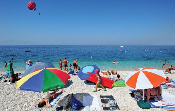 A beach with people sunbathing and swimming, colorful umbrellas, and boats in the water.