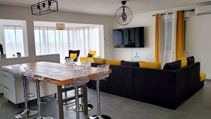 A modern living room with a black sofa, yellow cushions, a wooden dining table with metal chairs, a TV on the wall, and a ceiling light fixture.
