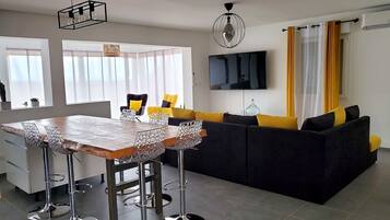 Una sala de estar moderna con un sofá negro, cojines amarillos, una mesa de comedor de madera con sillas metálicas, un televisor en la pared y una lámpara de techo.