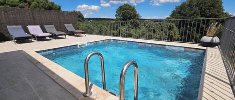 Une piscine rectangulaire avec des échelles en métal, entourée de chaises longues et d’une terrasse carrelée.