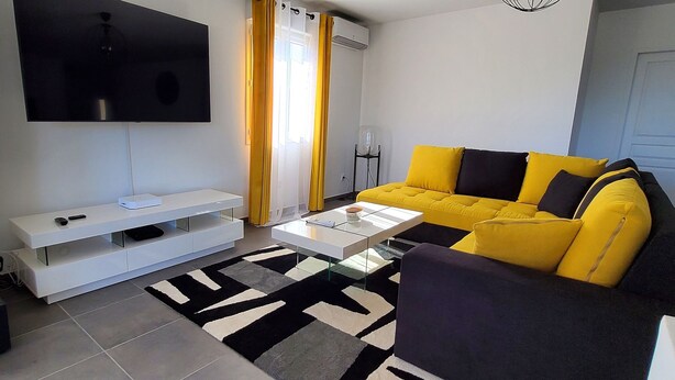 A modern living room with a yellow sofa, a flat-screen TV, a glass coffee table, and a black and white rug.