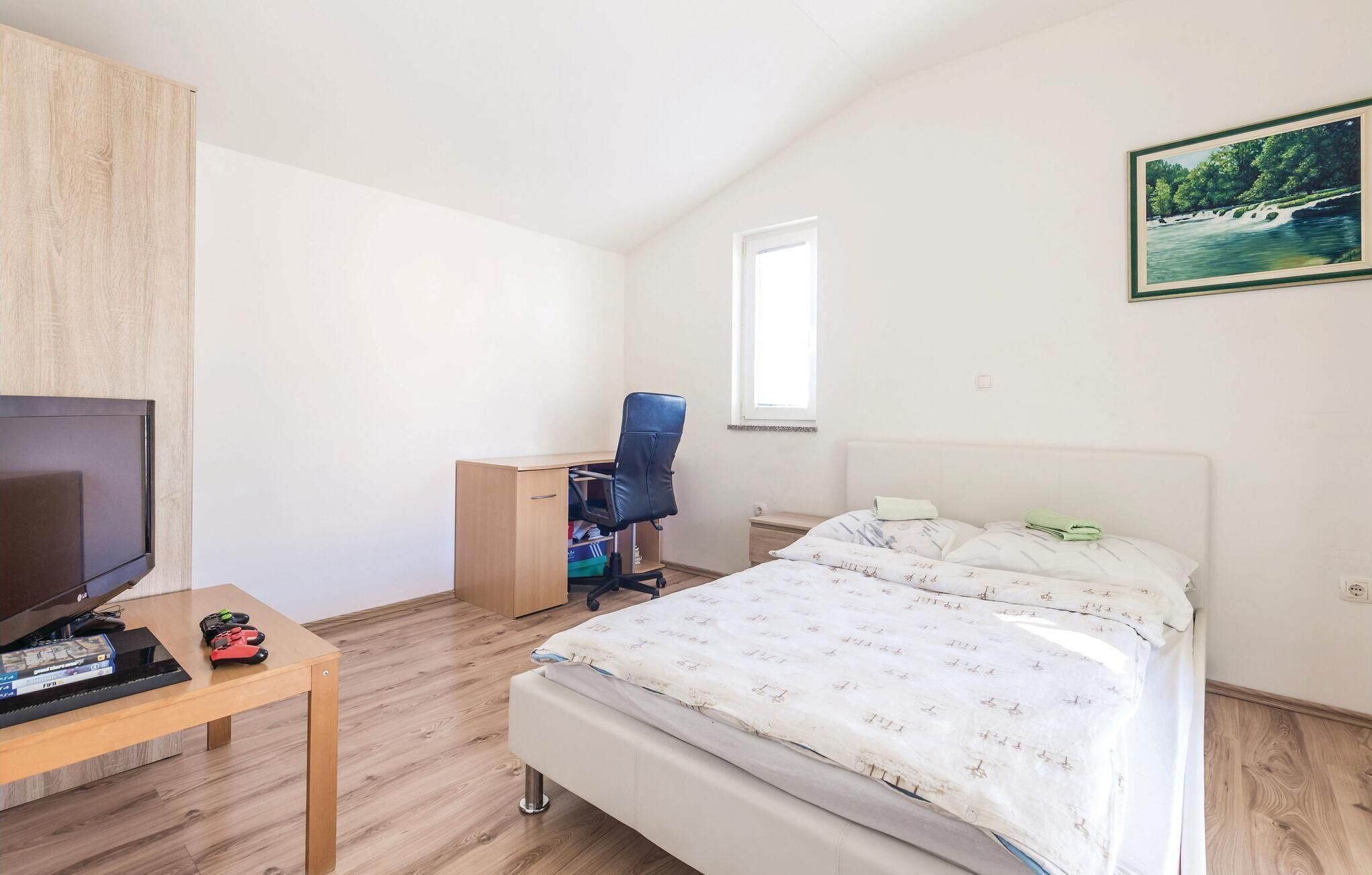 A bedroom with a bed, a desk with a chair, a television, and a window.