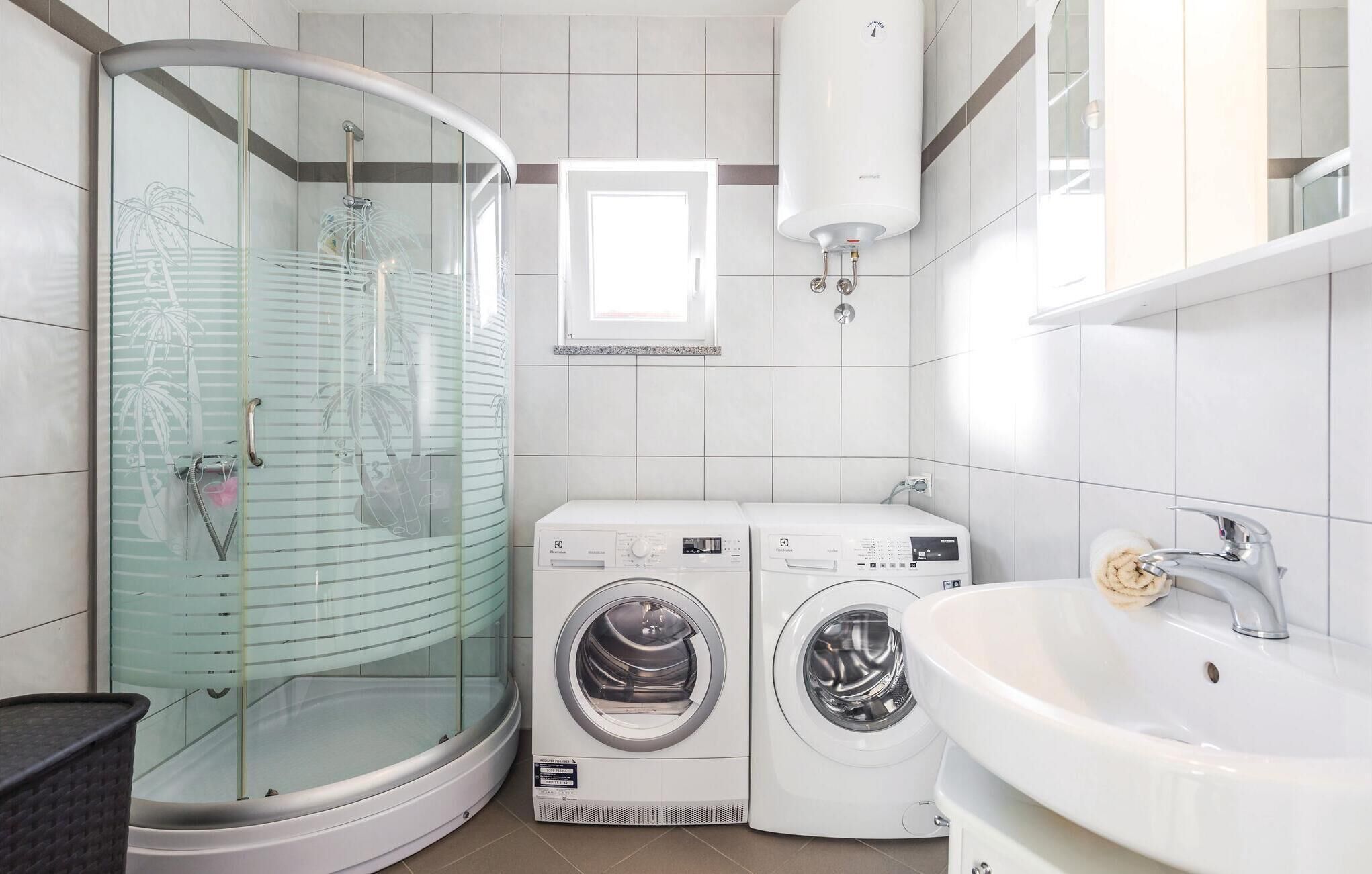 A modern bathroom with a glass shower enclosure, two front-loading washing machines, and a sink.