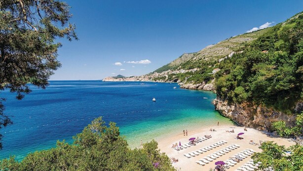 Ein Küstenstrand mit Reihen von Liegestühlen und Sonnenschirmen, einem klaren blauen Meer und einer hügeligen Kulisse.