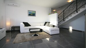 Un salón moderno con un sofá blanco, un otomano negro, una alfombra, un cuadro en la pared y una escalera con barandilla decorativa.