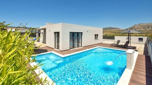 A modern outdoor pool area with a rectangular pool, a small building with large glass doors, and a view of a hilly landscape.