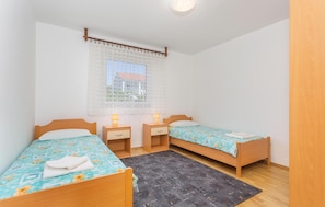 Quarto com duas camas de solteiro, janela com cortinas de renda, cabeceira de madeira e um tapete pequeno.