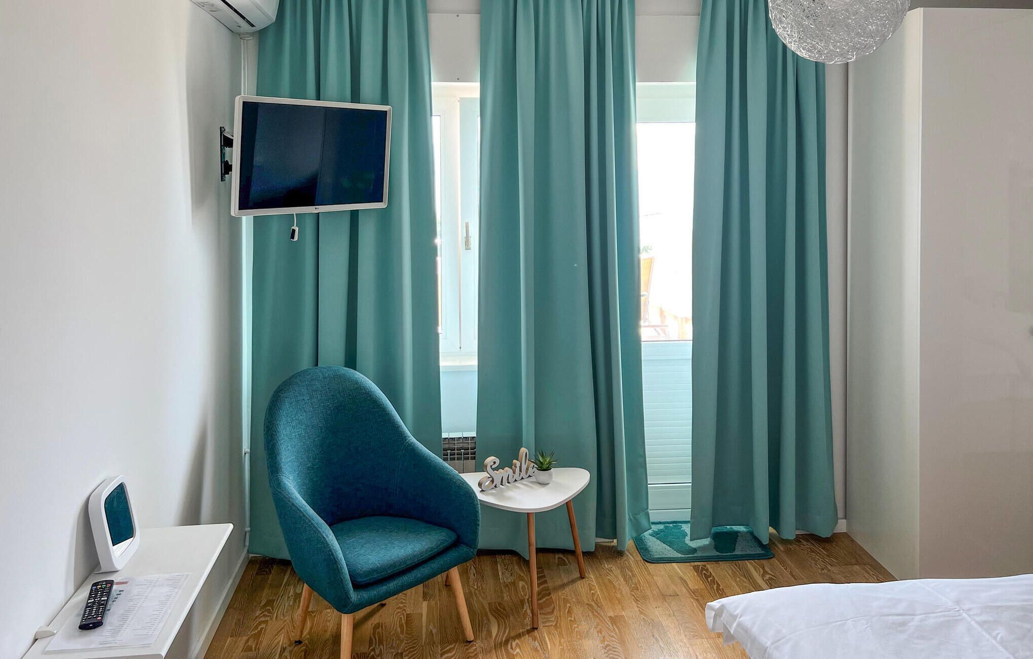 Habitación de hotel con cortinas verde marfil, un sillón verde marfil, una pequeña mesa blanca con una planta, un televisor en la pared y una cama con ropa de cama blanca.