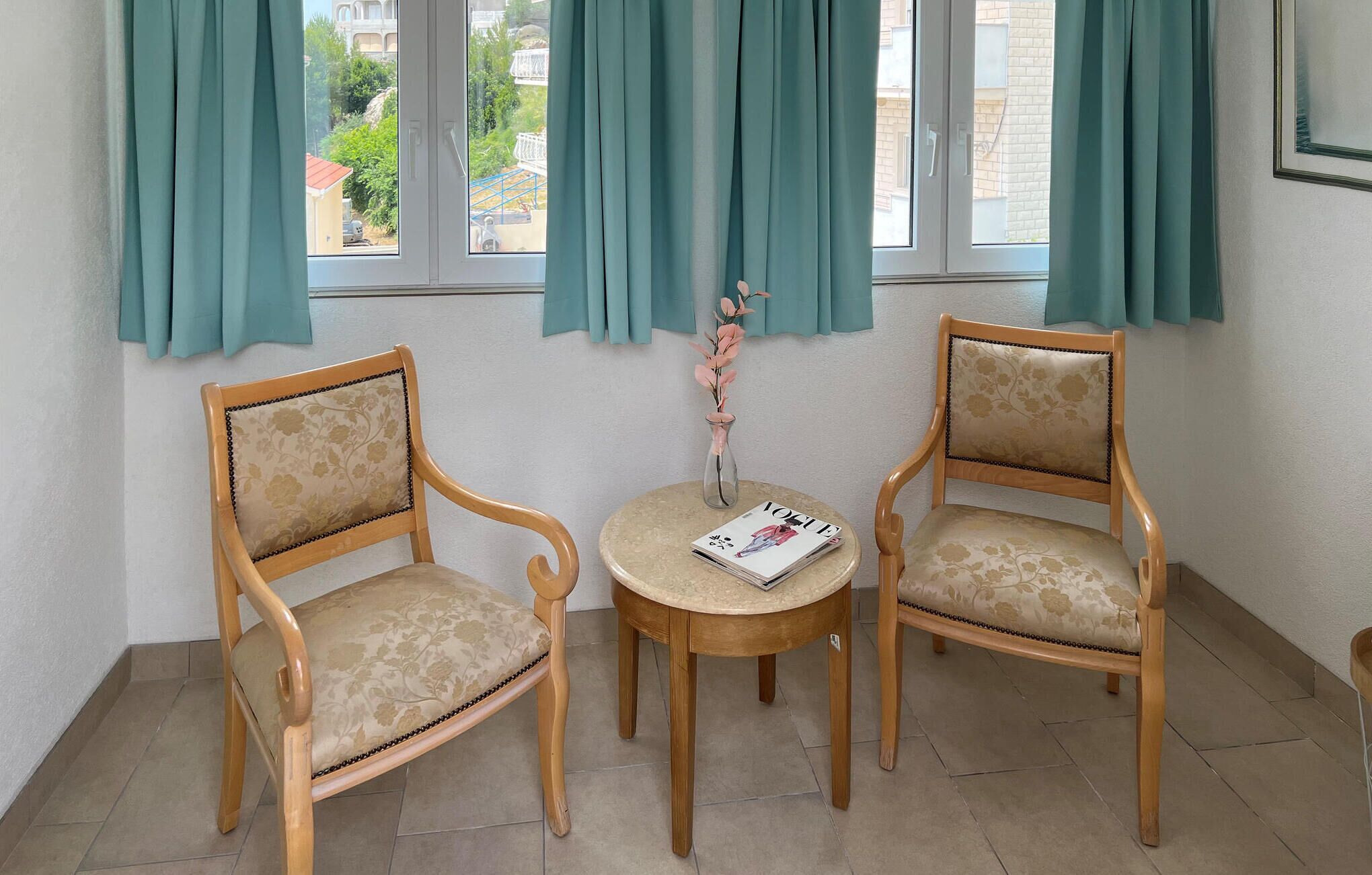 A room with two wooden chairs and a small round table, a vase with flowers, and teal curtains.