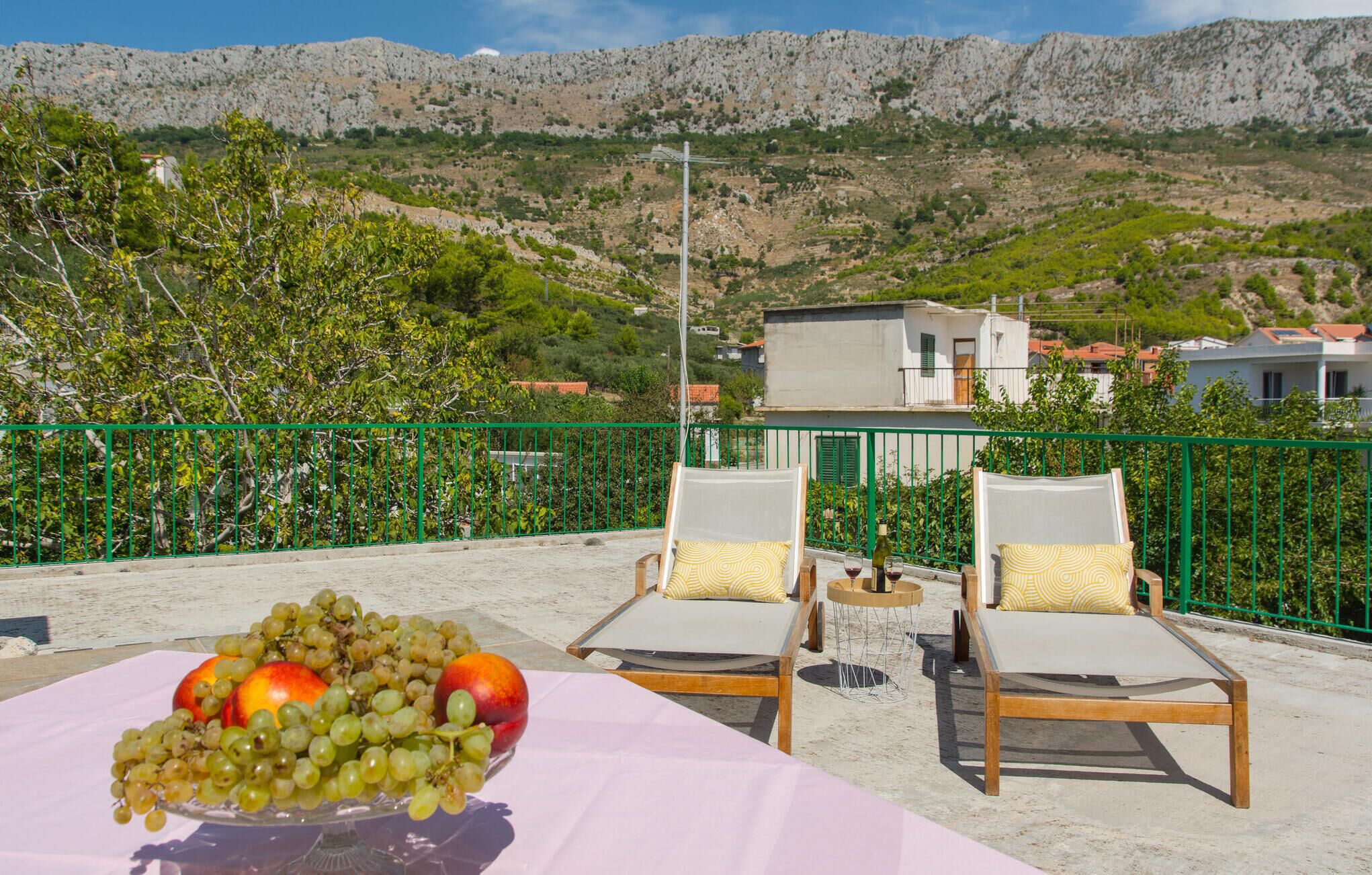 Une terrasse avec deux fauteuils, une table sur laquelle se trouve un panier de fruits, et une vue sur des collines rocheuses et des bâtiments.
