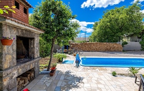 A stone fireplace with a hanging flower pot, a swimming pool, and a stack of firewood in the background. - Awesome Home in Drazice With Wifi and 2 Bedrooms (Jelenje)