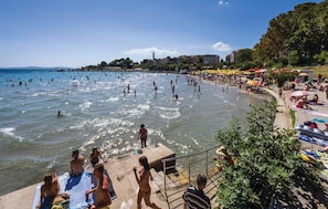 A crowded beach with people swimming and sunbathing, a pier with umbrellas, and buildings in the background. - Beautiful Home in Split With Wifi and 1 Bedrooms (Split)
