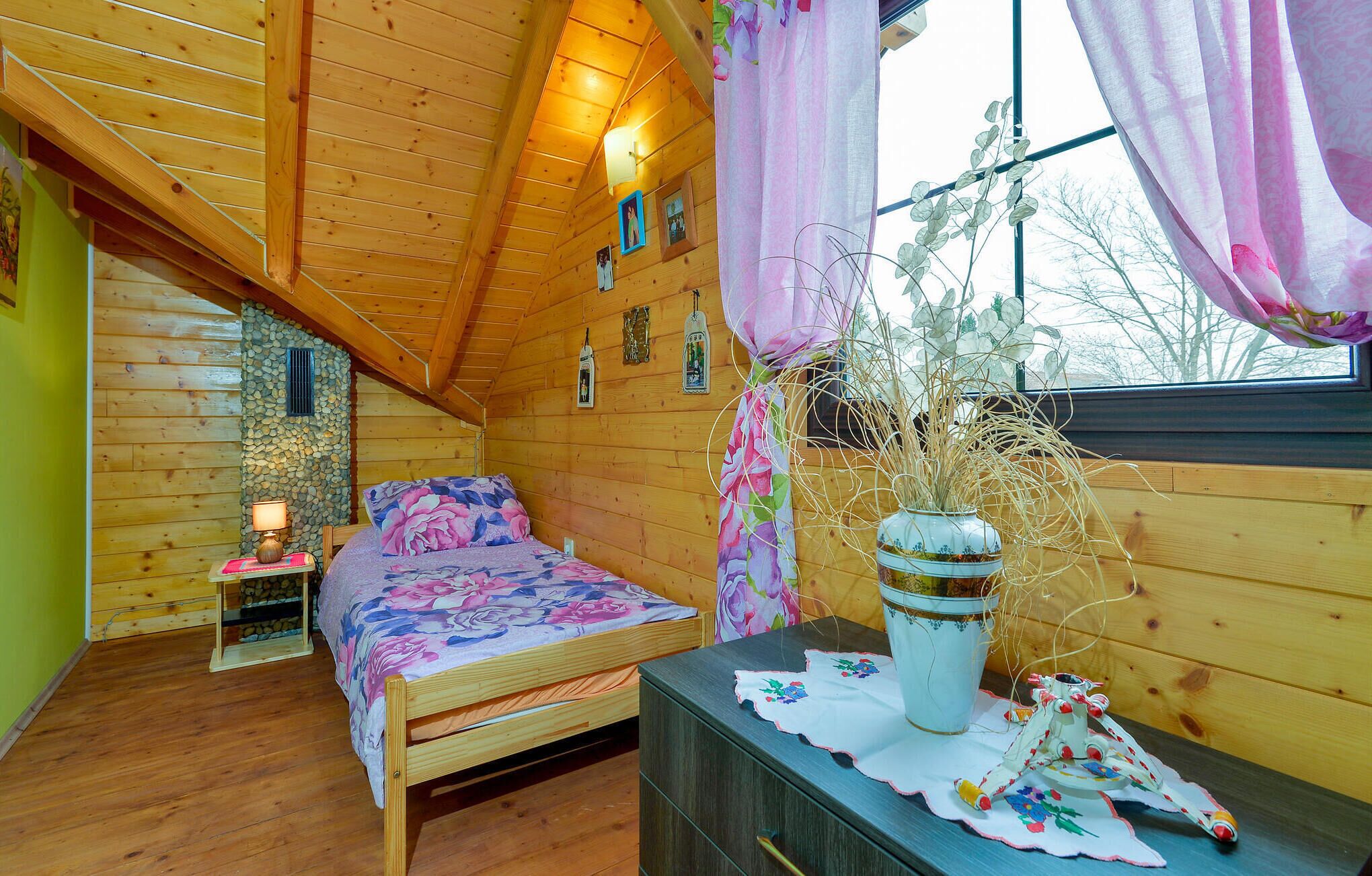 A cozy wooden cabin bedroom with a bed, a dresser, a vase with dried plants, and a window with curtains.