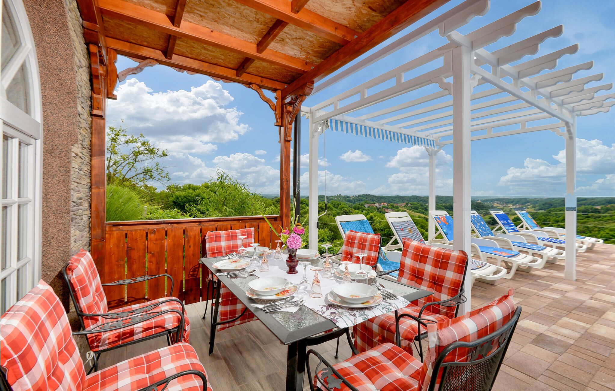 An outdoor dining area with a table set for four, surrounded by red and white checkered chairs. A white pergola provides shade, and a pool area with lounge chairs is visible in the background.