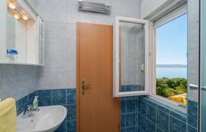 Ein Badezimmer mit Holztür, Waschbecken in Weiß, Spiegel und Fenster mit Meerblick.
