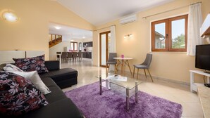 Ein modernes Wohnzimmer mit einer Couch, einem Glastisch und einem violetten Teppich. Im Hintergrund befindet sich ein Essbereich mit Stühlen und einem Tisch sowie eine Küche.
