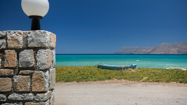 Un muro de piedra con una lámpara esférica, un sendero de tierra y un pequeño bote en un mar turquesa con montañas a la distancia.