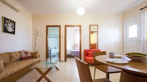 Una sala moderna con una mesa de centro de vidrio, una mesa de comedor de madera y un sofá. Hay una zona de cocina con una puerta blanca y un frutero sobre la mesa.