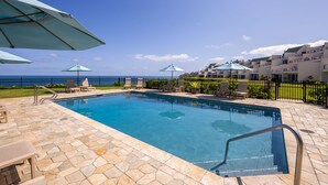 Piscina com espreguiçadeiras e guarda-sóis, cercada por um deck de pedra e uma grade.